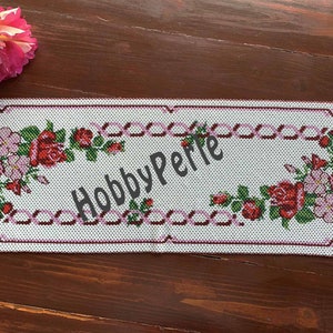 Può includere: Un runner da tavolo bianco ricamato con un motivo floreale rosa e bordeaux. Il motivo presenta rose e fiori rosa più piccoli. Il testo "HobbyPerle" è ricamato al centro del runner.