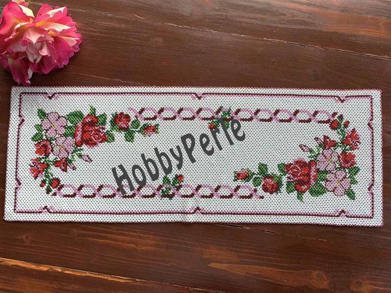 Pode incluir: Um corredor de mesa branco bordado com um design floral rosa e bord&ocirc;. O design apresenta rosas e flores rosa menores. O texto "HobbyPerle" &eacute; bordado no centro do corredor de mesa.