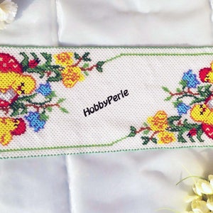 Può includere: Un runner da tavolo bianco perlato con un bordo verde e un motivo floreale con pulcini gialli, fiori rossi e foglie verdi. Il testo "HobbyPerle" è al centro del runner.