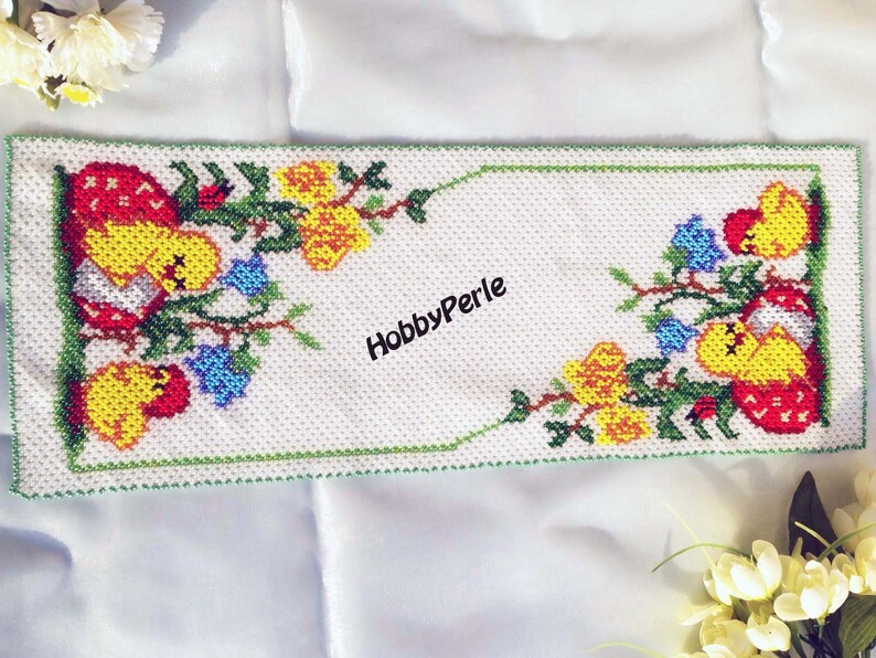 Pu&ograve; includere: Un runner da tavolo bianco perlato con un bordo verde e un motivo floreale con pulcini gialli, fiori rossi e foglie verdi. Il testo "HobbyPerle" &egrave; al centro del runner.