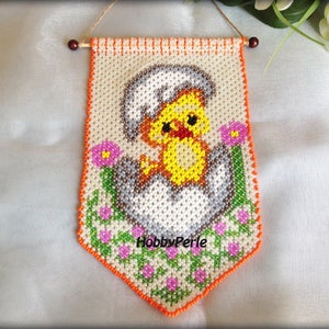 May include: A handmade Easter wall hanging featuring a yellow chick hatching from a white and grey egg. The chick is surrounded by pink flowers and green foliage. The banner is bordered with orange beads and has a wooden dowel for hanging.