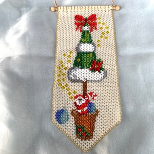 Può includere: Un'appendice da parete a forma di albero di Natale realizzata con perline, con un fiocco rosso, un albero verde, neve bianca e una figura di Babbo Natale in un vaso.