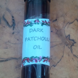 May include: A small glass bottle with a black cap and a white label that reads "DARK PATCHOULI OIL". The label has a red, green, and white floral design.