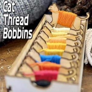 Op de afbeelding: Een houten doos met kattenvormige garenklossen. Elke klos bevat een andere kleur garen: rood, oranje, geel, groen, blauw en wit. De doos is gelabeld met "Cat Thread Bobbins".