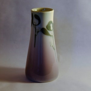 May include: A small, ceramic vase with a gradient of light purple to white. The vase has a floral design with green leaves and white flowers.