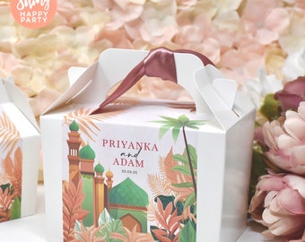 May include: White favor box with a pink ribbon handle. The box has a colorful design with a mosque, palm trees, and tropical leaves. The text on the box reads "Priyanka and Adam 28.09.25".