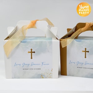 Puede incluir: Dos cajas de regalo blancas con detalles dorados y diseños de acuarela azul. Las cajas tienen una cruz dorada y el texto "Lucas George Dominic Fenner" y "SUNDAY 10TH OCTOBER".