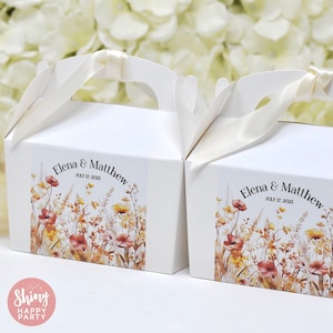 Op de afbeelding: Twee witte cadeauverpakkingen met handvatten en ivoorkleurig lint. Elke doos heeft een bloemenontwerp met rode, gele en bruine bloemen, en de tekst "Elena & Matthew JULY 17, 2025". Geschikt voor een bruiloft of feest.