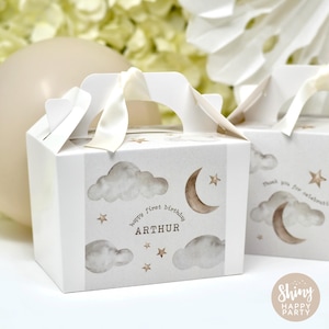 Puede incluir: Cajas de regalo blancas con asa y cinta color crema. Las cajas están decoradas con nubes de acuarela, lunas crecientes, estrellas y el texto "happy first birthday ARTHUR". Un globo y flores están en el fondo.