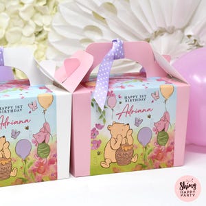 Pode incluir: Caixas rosa para lembrancinhas de festa com alças, com ilustrações de Winnie the Pooh, Piglet, balões e flores. As caixas são decoradas com o texto "Happy 1st Birthday Adriana". Uma fita roxa com bolinhas brancas está amarrada no topo.