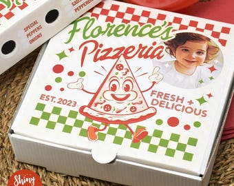 May include: White pizza box with a red, green, and white checkered pattern. The box has a cartoon pizza character with a smiling face and the text "Florence's Pizzeria" in a red and green font. The text "Fresh + Delicious" and "Est. 2023" are also on the box. A photo of a child is in a circle on the box.
