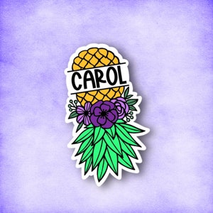 May include: A sticker featuring a pineapple with the name "CAROL" written on it. The pineapple is surrounded by green leaves and purple flowers.