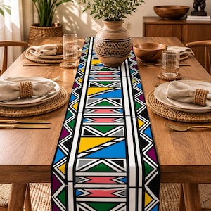 Puede incluir: Un camino de mesa rectangular largo con un patrón geométrico audaz de triángulos y rayas repetitivas en colores vibrantes que incluyen amarillo, azul, verde, rosa, morado, gris, negro y blanco, centrado sobre una mesa de comedor de madera.
