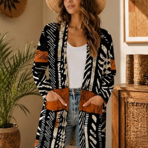 Op de afbeelding: Een vest met lange mouwen en een open voorzijde, voorzien van een abstract geometrisch patroon in zwart, wit en gebrand oranje. Het kledingstuk heeft twee opgestikte zakken aan de voorzijde en een rechte zoom, gedragen over een wit hemdje.