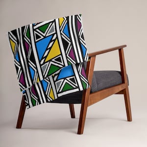 May include: A colorful throw blanket with a geometric pattern in shades of black, white, yellow, blue, green, and purple. The blanket is folded and draped over a wooden chair.
