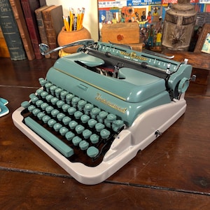 May include: A teal and cream vintage Underwood typewriter. The typewriter has a full keyboard with teal keys, a chrome carriage return lever, and the Underwood logo in gold script. It is placed on a wooden surface.