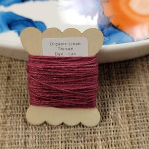 May include: A wooden spool of organic linen thread dyed a deep burgundy color. The spool has a scalloped edge and a small label that reads "Organic Linen Thread Dye - Lac". The thread is tightly wound around the spool.
