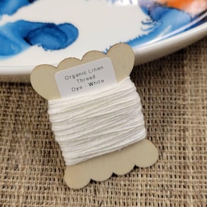 May include: A spool of white organic linen thread labeled "Organic Linen Thread Dye - White." The thread is wound around a light brown, decorative spool. The background includes a white plate with blue and orange paint and a brown woven surface.