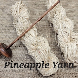May include: Two skeins of natural-colored pineapple yarn, a traditional fiber, are displayed on a woven burlap background. A wooden spinning wheel is also shown.