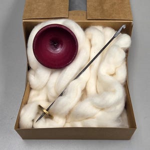 May include: A craft kit in a cardboard box, featuring a deep red wooden bowl, a black spindle with a gold whorl, and a mass of fluffy, off-white wool roving. The kit is designed for spinning yarn.
