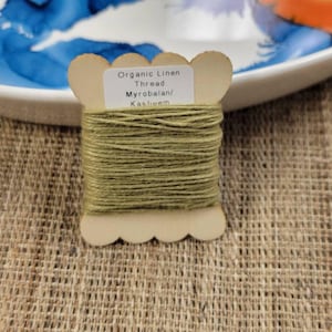 May include: A spool of olive green organic linen thread labeled "Myrobalan/Kasheem" sits on a textured surface. The thread is wound around a light-colored wooden spool with a scalloped edge. The background includes a white plate with blue and orange paint.