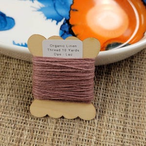 May include: A wooden spool of organic linen thread in a muted mauve color. The label reads "Organic Linen Thread 10 Yards Dye - Lac". The spool is set against a textured, neutral background, with a decorative plate in the background.