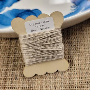 May include: A spool of natural-colored organic linen thread. The thread is wound around a light brown, scalloped-edge cardboard spool. A small white label on the spool reads "Organic Linen Thread, Dye - Natural". The spool sits on a textured, tan surface.