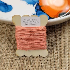 May include: A spool of organic linen thread dyed madder, with a label that reads "Organic Linen Thread 10 Yards Dye - Madder." The thread is a warm coral color, wound around a wooden spool. The background includes a white plate with blue and orange paint.