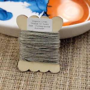 May include: A wooden spool of organic linen thread, dyed with pomegranate and kasheem. The thread is a muted grey colour and is labelled as 9.14 metres. The spool has a decorative, scalloped edge.