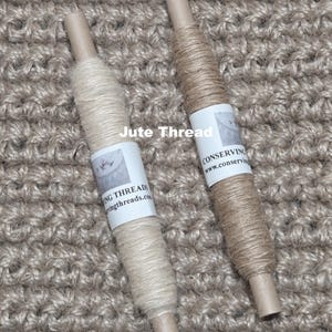 May include: Two spools of jute thread, one light beige and one tan, are shown against a textured, woven background. Each spool has a paper label with text. The words "Jute Thread" are printed across the top of the image.