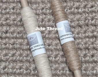 Puede incluir: Dos carretes de hilo de yute, uno beige claro y otro marrón claro, se muestran sobre un fondo texturizado y tejido. Cada carrete tiene una etiqueta de papel con texto. Las palabras "Jute Thread" están impresas en la parte superior de la imagen.