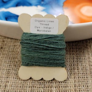 May include: A wooden spool of organic linen thread dyed indigo and myrobalan. The thread is a deep forest green color and is wrapped around the spool. The label on the spool reads "Organic Linen Thread Dye - Indigo / Myrobalan."