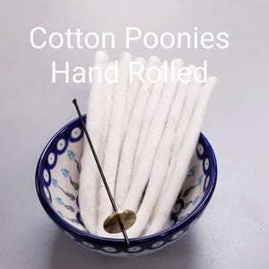 May include: A ceramic bowl with a blue and white floral pattern holds a bundle of white cotton wicks. A black and brass wick dipper is placed in the bowl. The text "Cotton Poonies Hand Rolled" is displayed above the bowl.