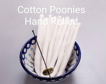 May include: A ceramic bowl with a blue and white floral pattern holds a bundle of white cotton wicks. A black and brass wick dipper is placed in the bowl. The text "Cotton Poonies Hand Rolled" is displayed above the bowl.