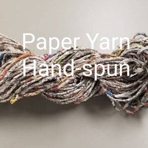 May include: A skein of hand-spun paper yarn. The yarn is made from recycled paper and has a natural, rustic look. The yarn is a mix of white, brown, and beige colours with hints of blue, red, and yellow.