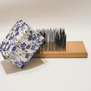 Puede incluir: Un kenzan de madera con alfileres metálicos afilados, junto a un recipiente cuadrado con un estampado floral azul sobre fondo blanco. El recipiente está abierto e inclinado.