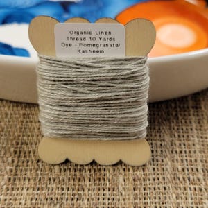 May include: A wooden spool of organic linen thread, dyed in a neutral grey. The label reads "Organic Linen Thread 10 Yards Dye - Pomegranate/ Kasheem". The thread is wound around a decorative wooden spool. The background includes a white bowl and a textured surface.