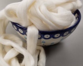 May include: A ceramic bowl with a blue rim and white and blue floral designs is filled with fluffy, off-white wool roving. Strands of the wool spill out of the bowl, creating a soft, textured display.