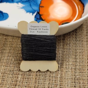 May include: A wooden spool of black organic linen thread, approximately 9.1 metres, with the text "Organic Linen Thread 10 Yards Dye - Kasheem". The spool is set against a textured, neutral-toned background, with a decorative bowl in the background.