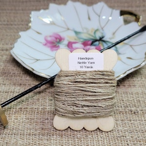 May include: A spool of handspun nettle yarn with a label that reads "Handspun Nettle Yarn 10 Yards." A black spindle and a decorative leaf-shaped dish are in the background. The yarn is a natural beige colour.