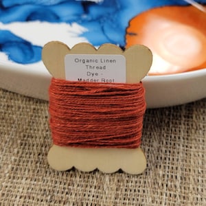 May include: A spool of organic linen thread dyed with madder root. The thread is a rich, reddish-brown color and is wound around a light brown, decorative spool. The label on the spool reads "Organic Linen Thread Dye - Madder Root".