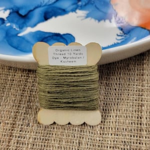May include: A spool of organic linen thread dyed with myrobalan and kasheem. The thread is a muted olive green color and is wound around a wooden spool. The label reads "Organic Linen Thread 10 Yards".
