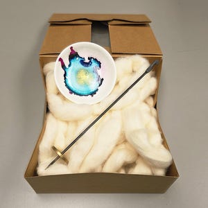 May include: A craft kit in a cardboard box, containing white wool roving, a small ceramic bowl with a colorful abstract design, and a black needle tool. The bowl features blue, yellow, and pink hues.