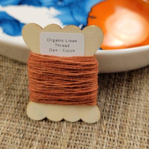 May include: A spool of organic linen thread dyed with cutch. The thread is a warm, earthy orange color, wound around a wooden spool with a scalloped edge. The label reads "Organic Linen Thread Dye - Cutch."