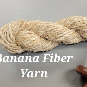 May include: A close-up of a skein of banana fibre yarn, a natural beige colour, with a wooden spindle. The text "Banana Fiber Yarn" is displayed in white. This yarn is suitable for various crafting projects.