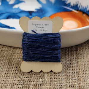 May include: A wooden spool of organic linen thread dyed indigo. The spool has a scalloped edge and a label that reads "Organic Linen Thread Dye - Indigo". The thread is a deep navy blue color. The background includes a white bowl with blue and orange accents.