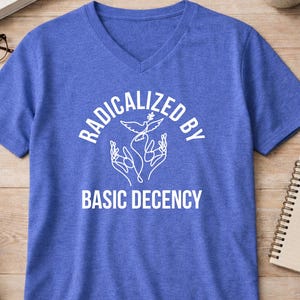 Peut inclure: Un t-shirt bleu à col en V avec le texte blanc "RADICALIZED BY BASIC DECENCY". Le design comprend une colombe et des mains. Le t-shirt est en matière souple. Vêtement décontracté.