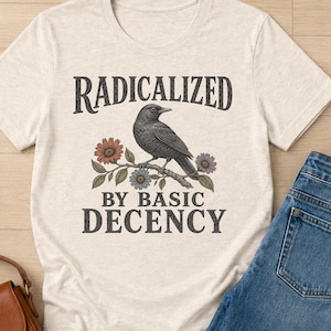 Może przedstawiać: Kremowy t-shirt z krótkim rękawem z czarną grafiką przedstawiającą ptaka siedzącego na kwiecistej gałęzi. Napis RADICALIZED BY BASIC DECENCY jest nadrukowany czarną czcionką szeryfową powyżej i poniżej ilustracji ptaka.
