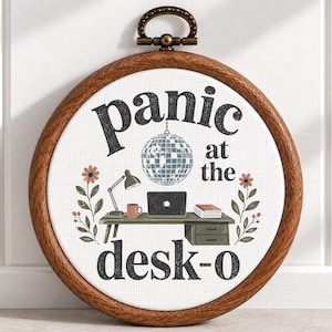 May include: A circular wooden embroidery hoop containing white fabric with the text "panic at the desk-o" in black. The design features a silver disco ball, a desk with a laptop, a lamp, a mug, a stack of books, and two floral stems.