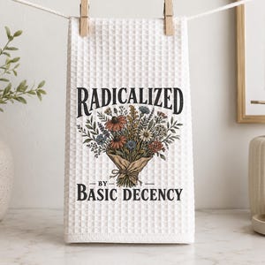 May include: A white waffle-weave kitchen towel hanging from a clothesline with two wooden clips. The towel features a central graphic of a colorful wildflower bouquet wrapped in paper, with the text "RADICALIZED BY BASIC DECENCY" printed in black.